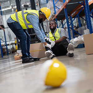 Worker helping injured colleague on warehouse floor, symbolizing workplace safety and injury.